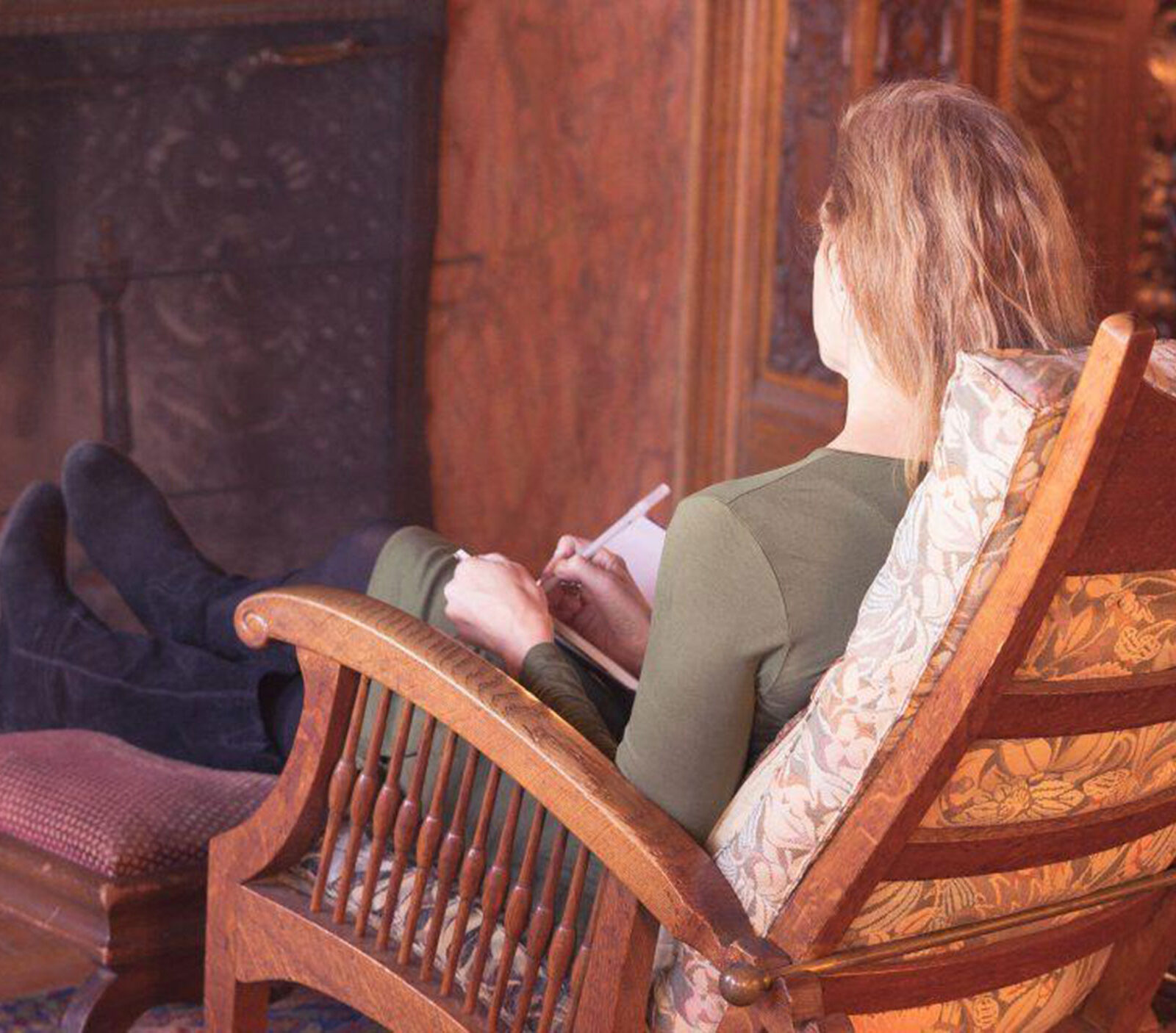 person sits in a wooden chairs, feet up in front of a fireplace; writes in a notebook