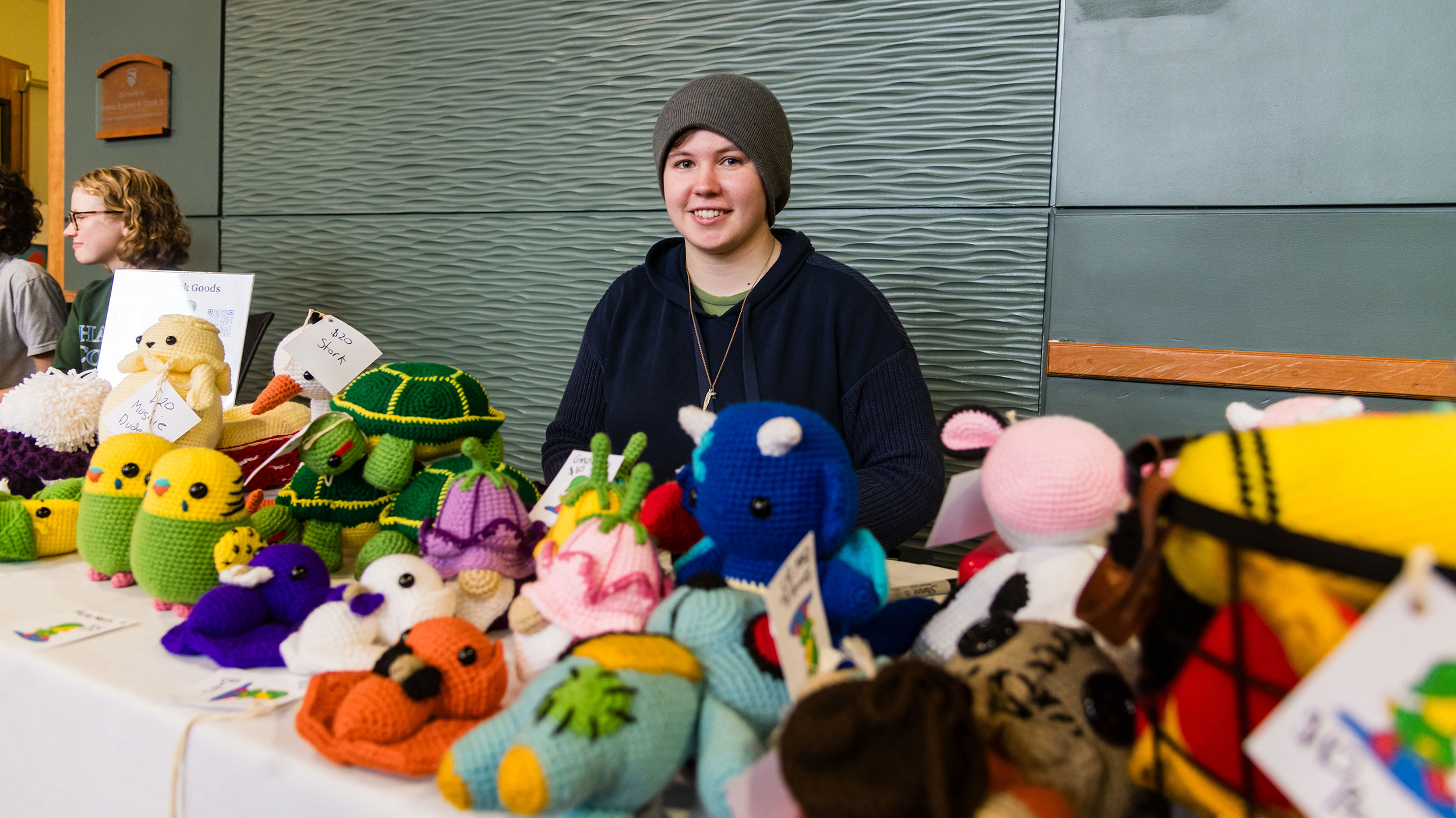 a student sits at a table covered in homemade crocheted plushies that they are selling