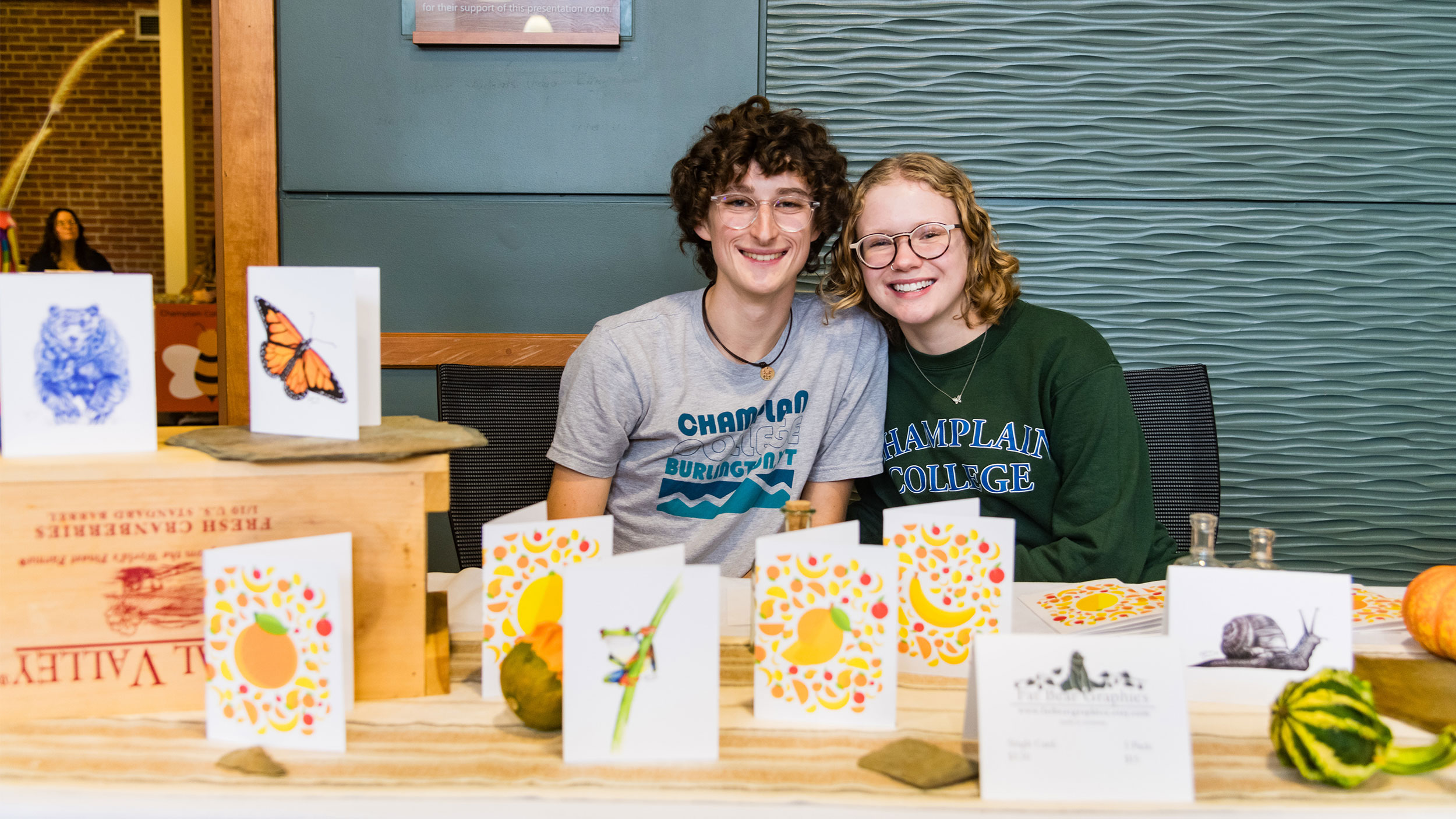 two students smile big sitting at a table covered in homemade cards with yellow fruit drawn on them