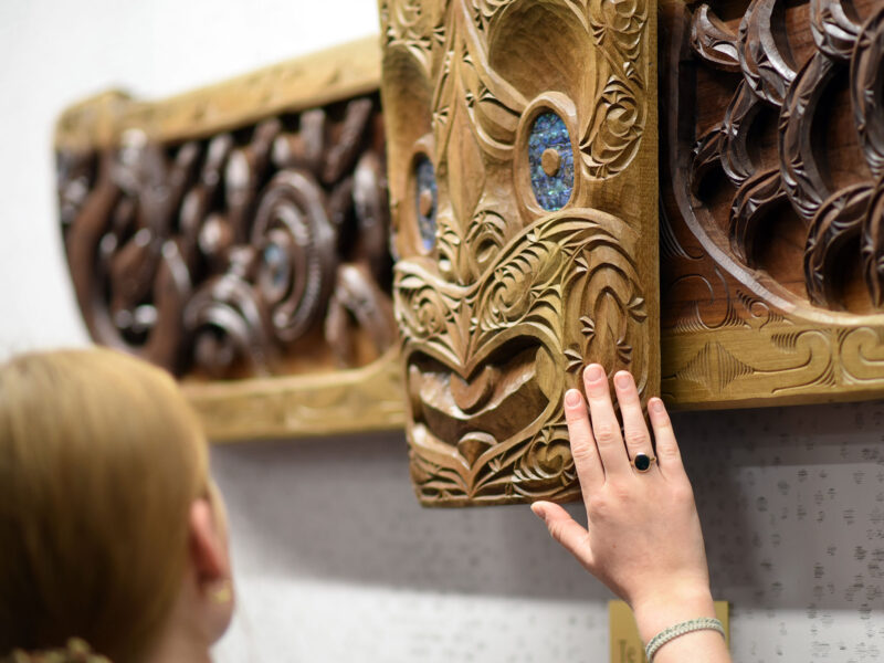 a student reaches their hand to a wood carving on the wall by a maori artist