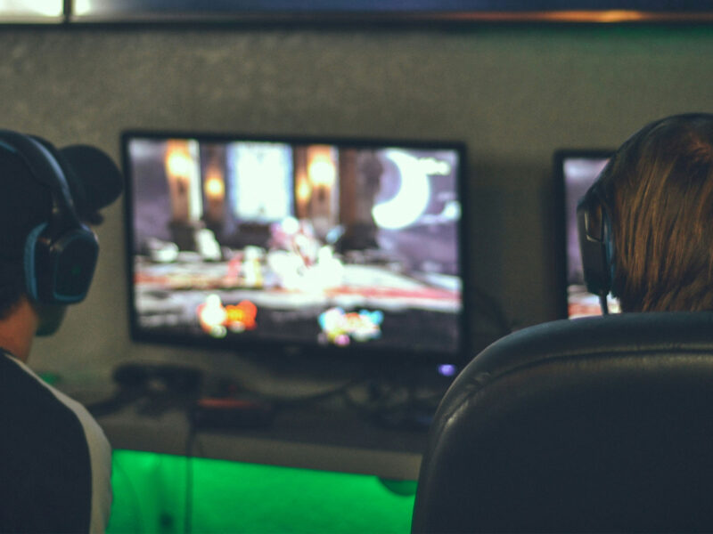 view from the back of two people sitting down gaming, with a blurred computer screen in the distance