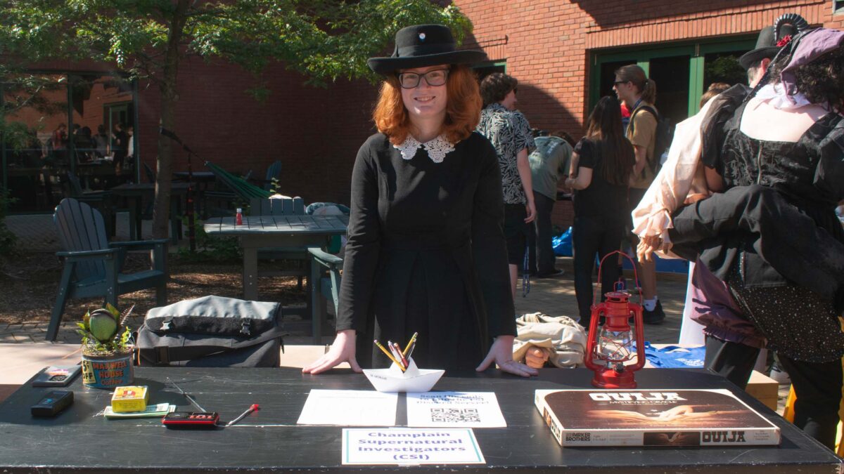 a student standing behind the Champlain CSI club table with a red lantern, ouija board, and various horror movie memorabilia at the activity fair