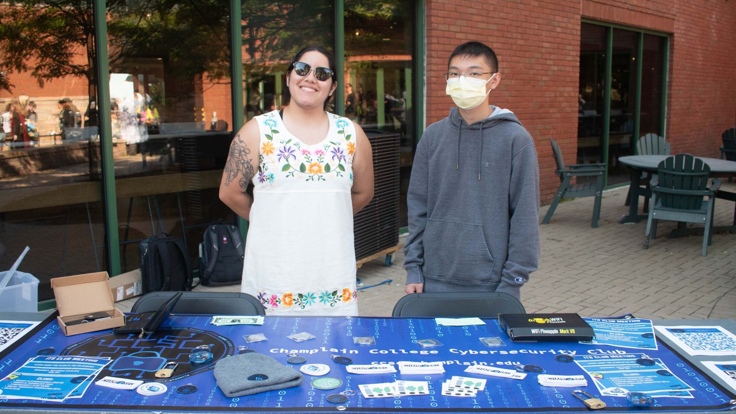 two students standing behind the Champlain College Cybersecurity Club table at the activity fair in the courtyard on campus