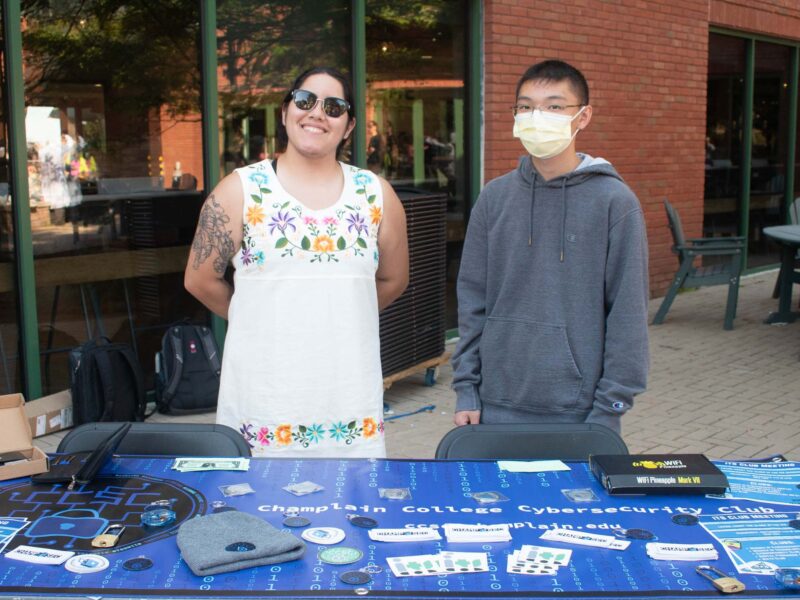 two students standing behind the Champlain College Cybersecurity Club table at the activity fair in the courtyard on campus