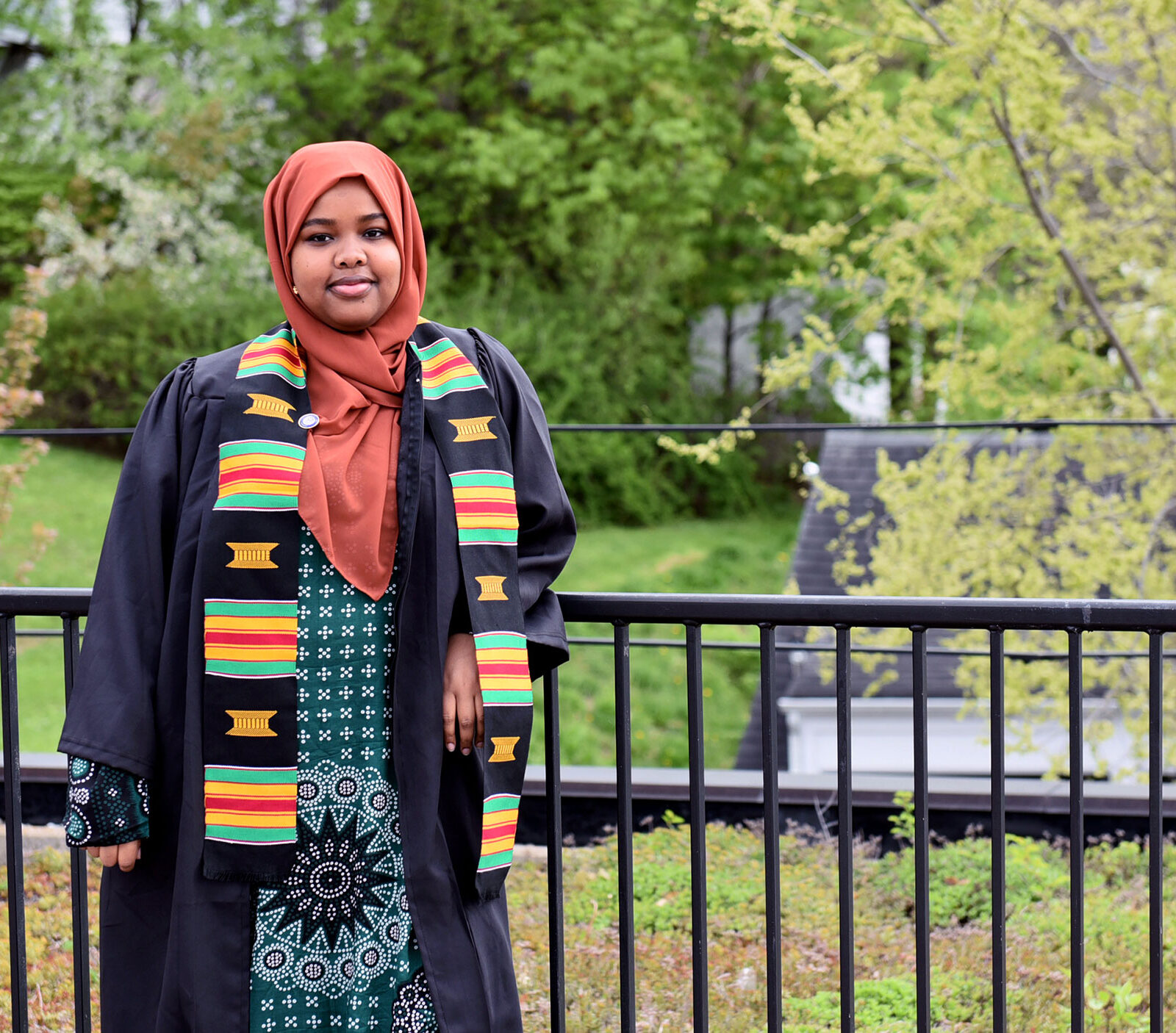 Hawa Adan wearing a graduation gown and ODI sash standing outside leaning against a black railing