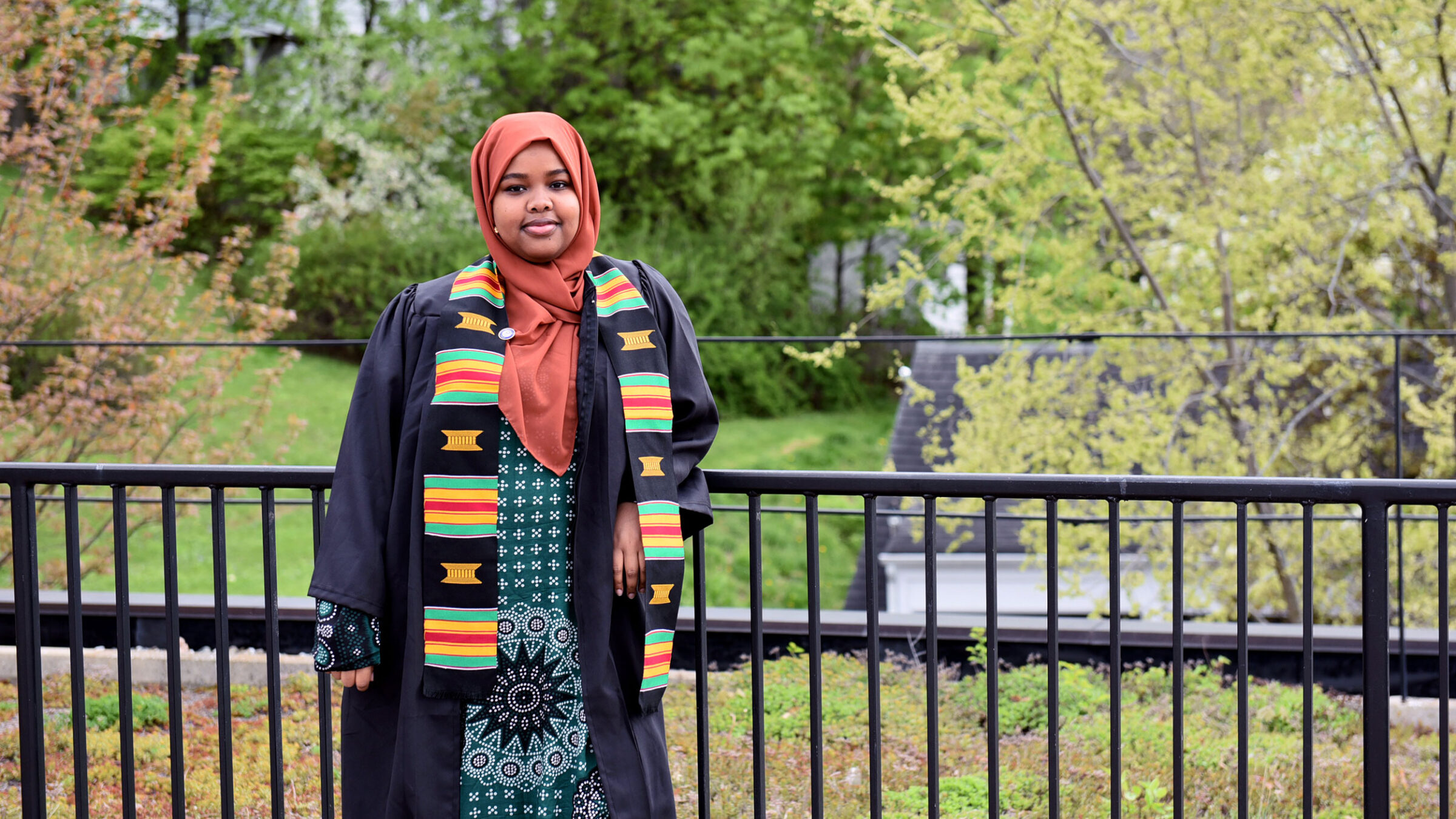 Hawa Adan wearing a graduation gown and ODI sash standing outside leaning against a black railing