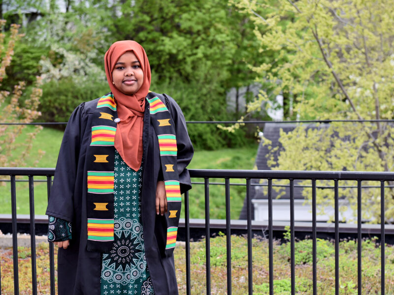 Hawa Adan wearing a graduation gown and ODI sash standing outside leaning against a black railing