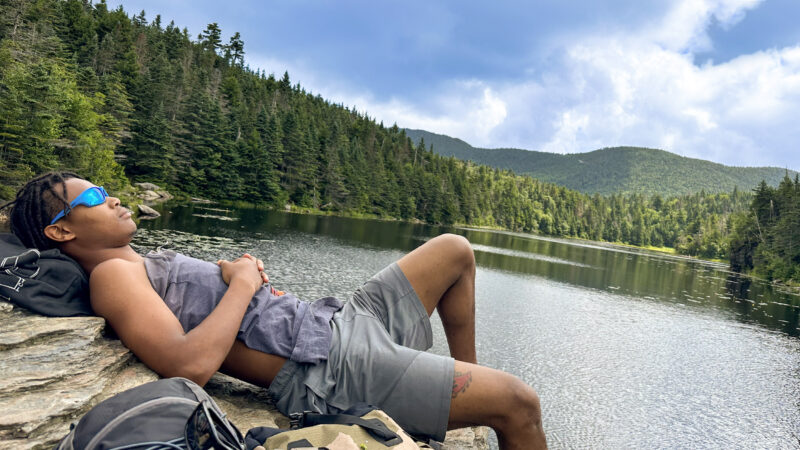 student rests on a rock by a lake after a hike