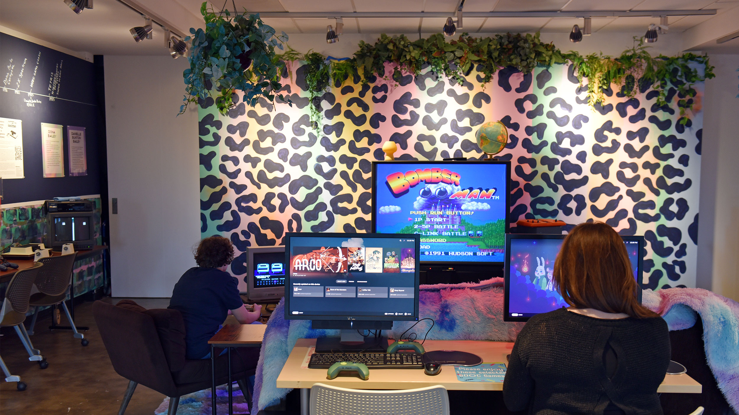 two people play games on a PC and game console in front of a big colorful wall