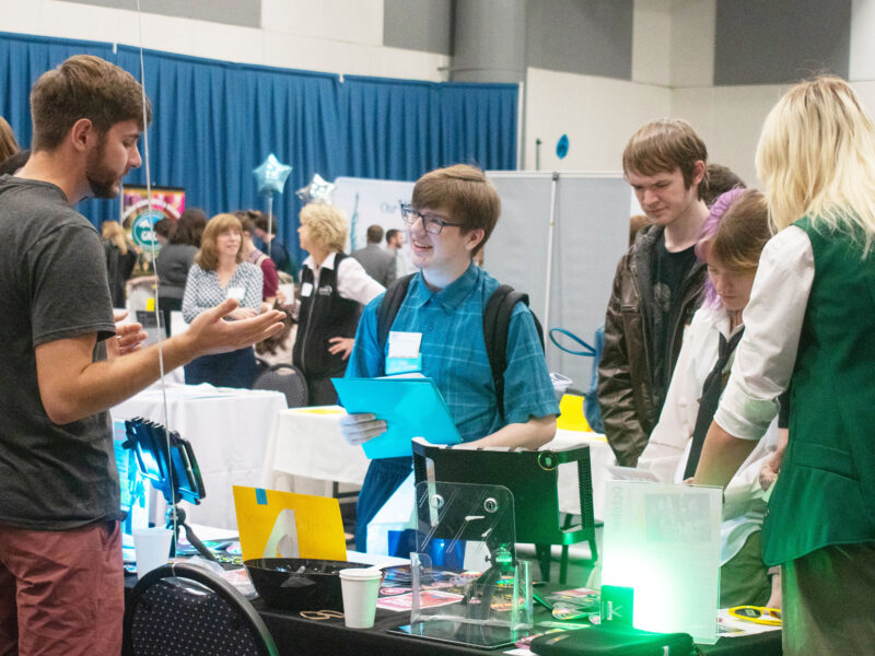 Students look at an employer's table display at the Career Fair and listen closely to the employer speaking