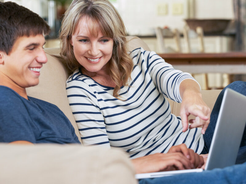 Mother With Teenage Son Sitting On Sofa At Home Using Laptop