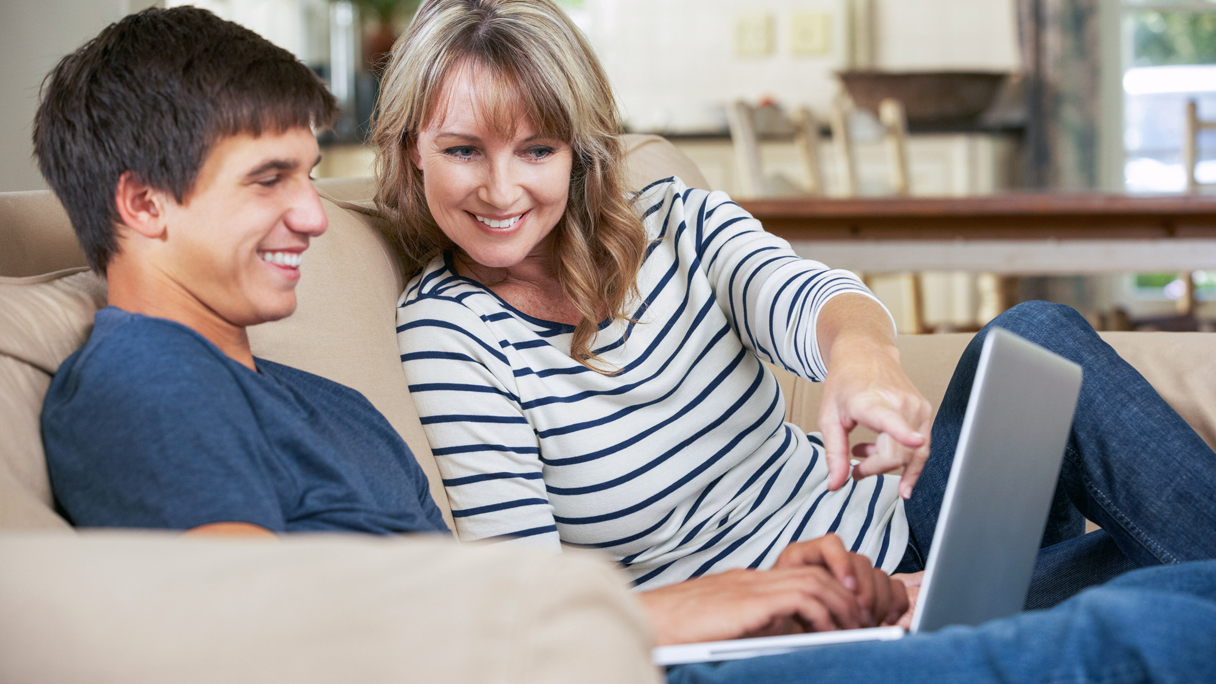 Mother With Teenage Son Sitting On Sofa At Home Using Laptop