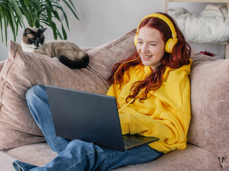 Smiling young Caucasian woman sit on couch in living room typing working on laptop, happy millennial teen girl browsing wireless internet, texting messaging on computer at home