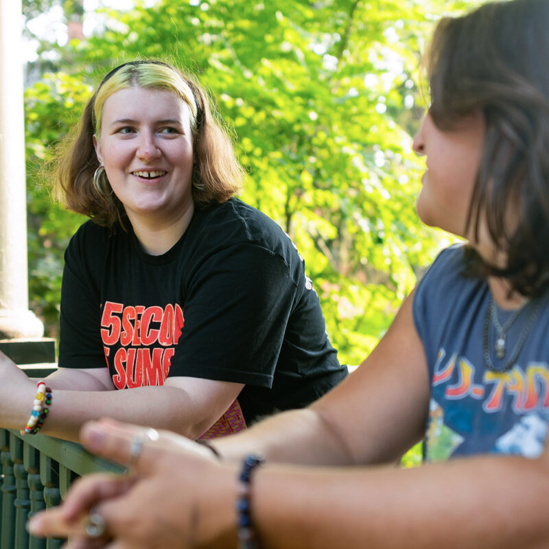 two students talk while standing on the front porch of a residence hall