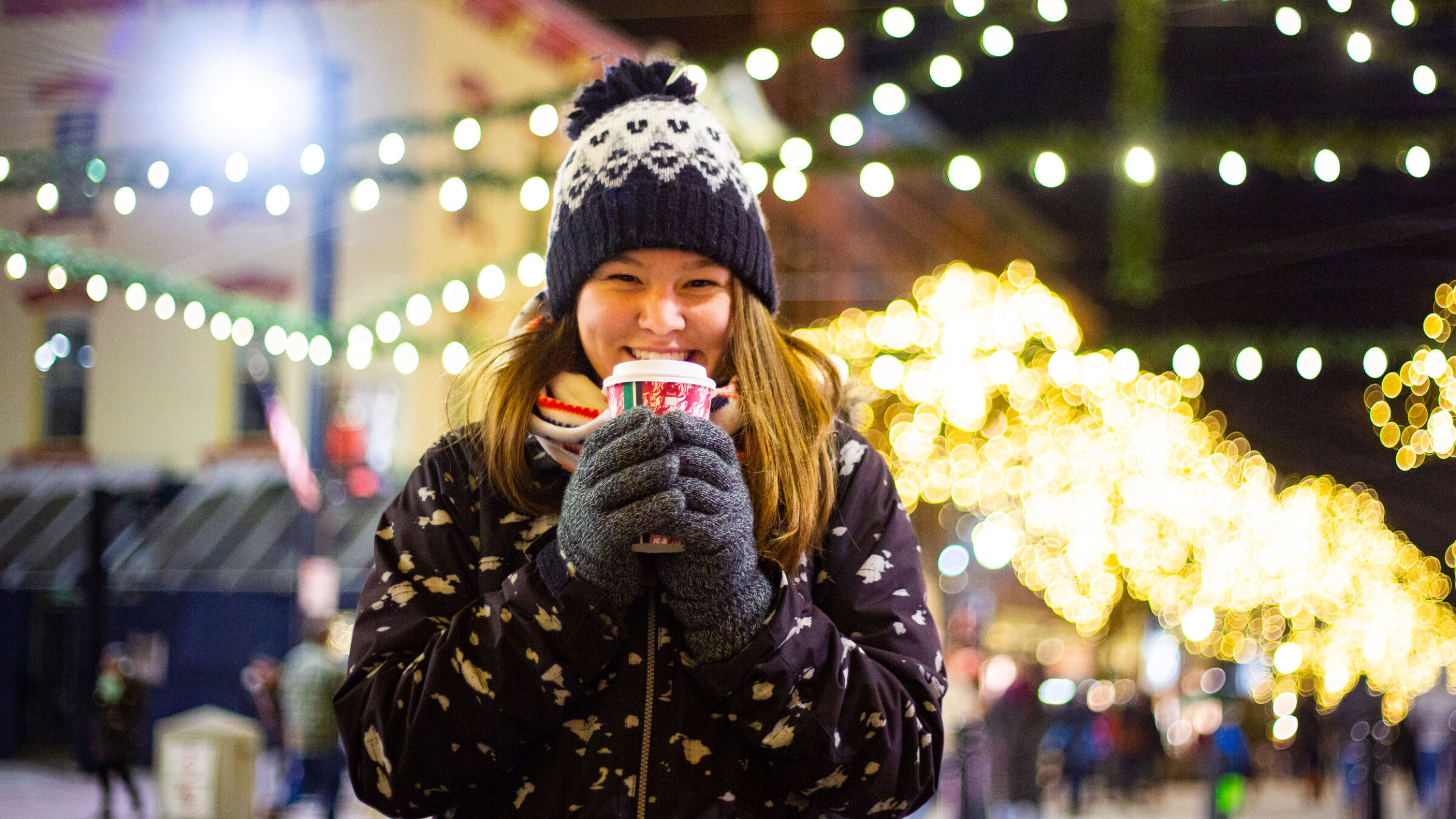 Student holding a cup of hot chocolate on Church Street in the winter, surrounded by Christmas lights