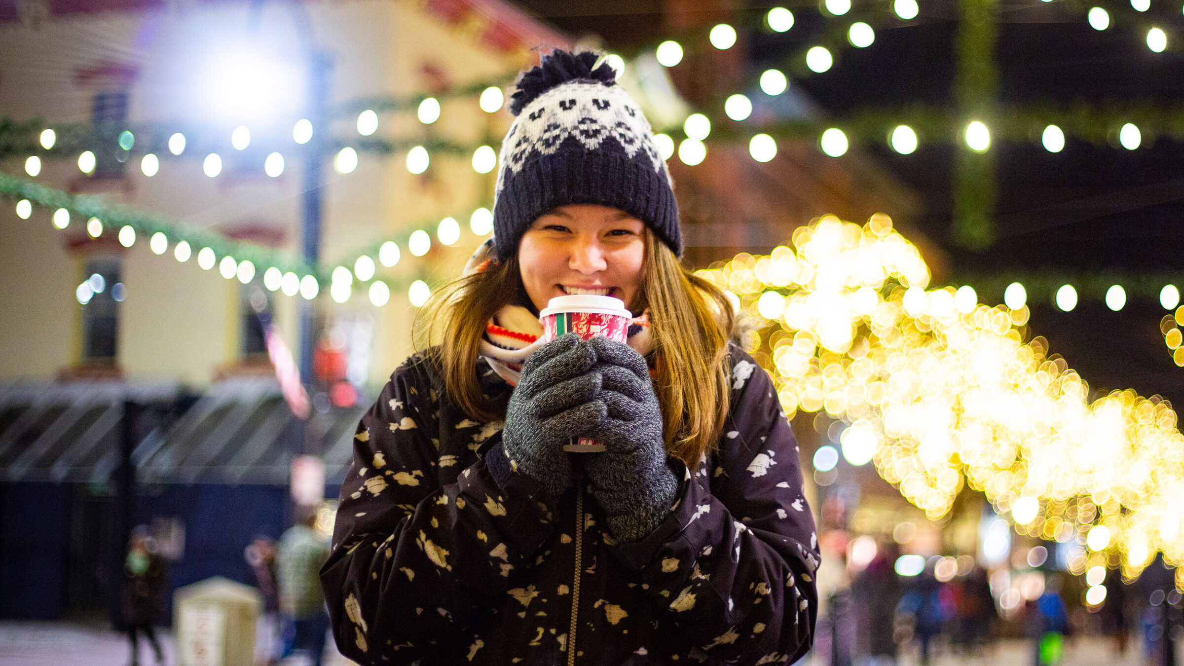 Student holding a cup of hot chocolate on Church Street in the winter, surrounded by Christmas lights