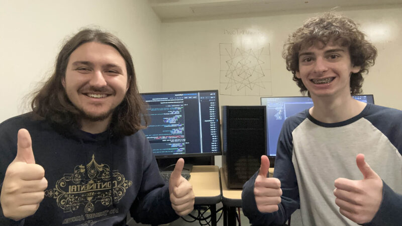 Two students smiling and giving a thumbs up in front of a computer in a classroom